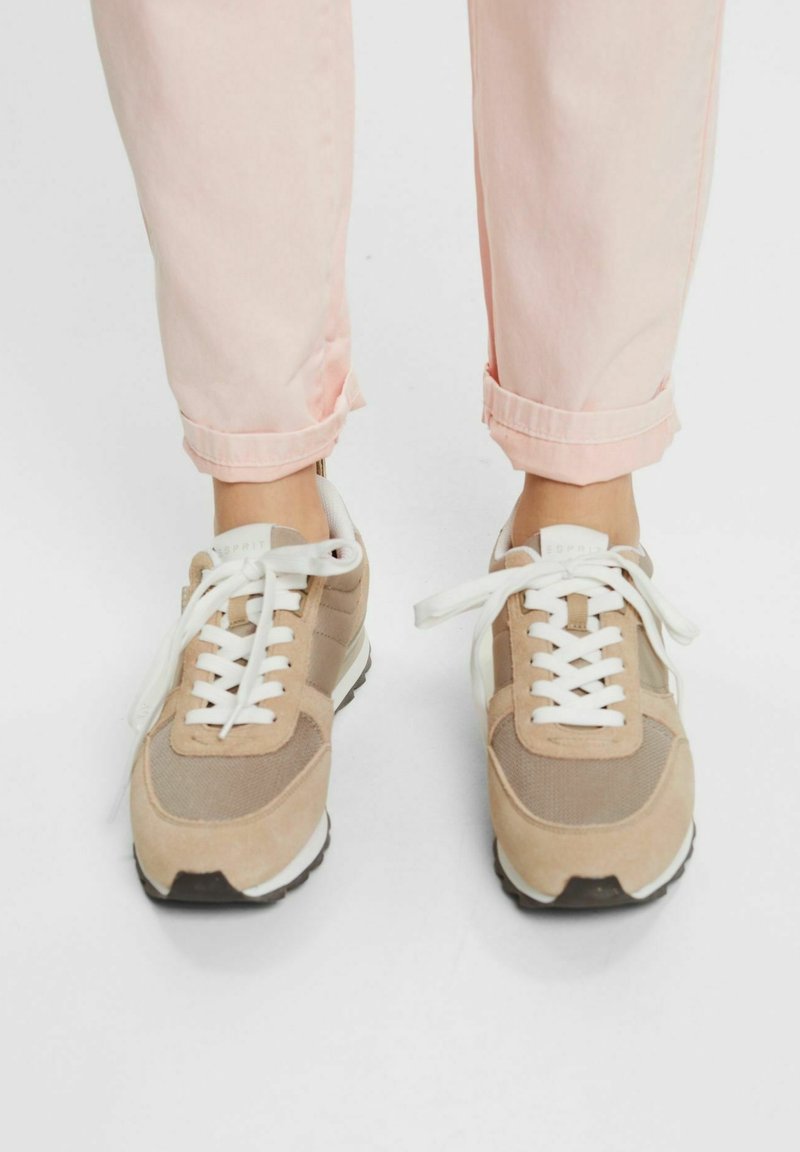 Pieds portant des baskets beiges et blanches à lacets assorties à un pantalon rose clair retroussé sur un fond blanc uni.