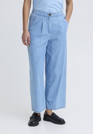 Personne portant un pantalon en denim bleu clair à jambes larges avec des plis devant, un haut bleu clair, des mocassins en cuir noir et des bagues à la main gauche.