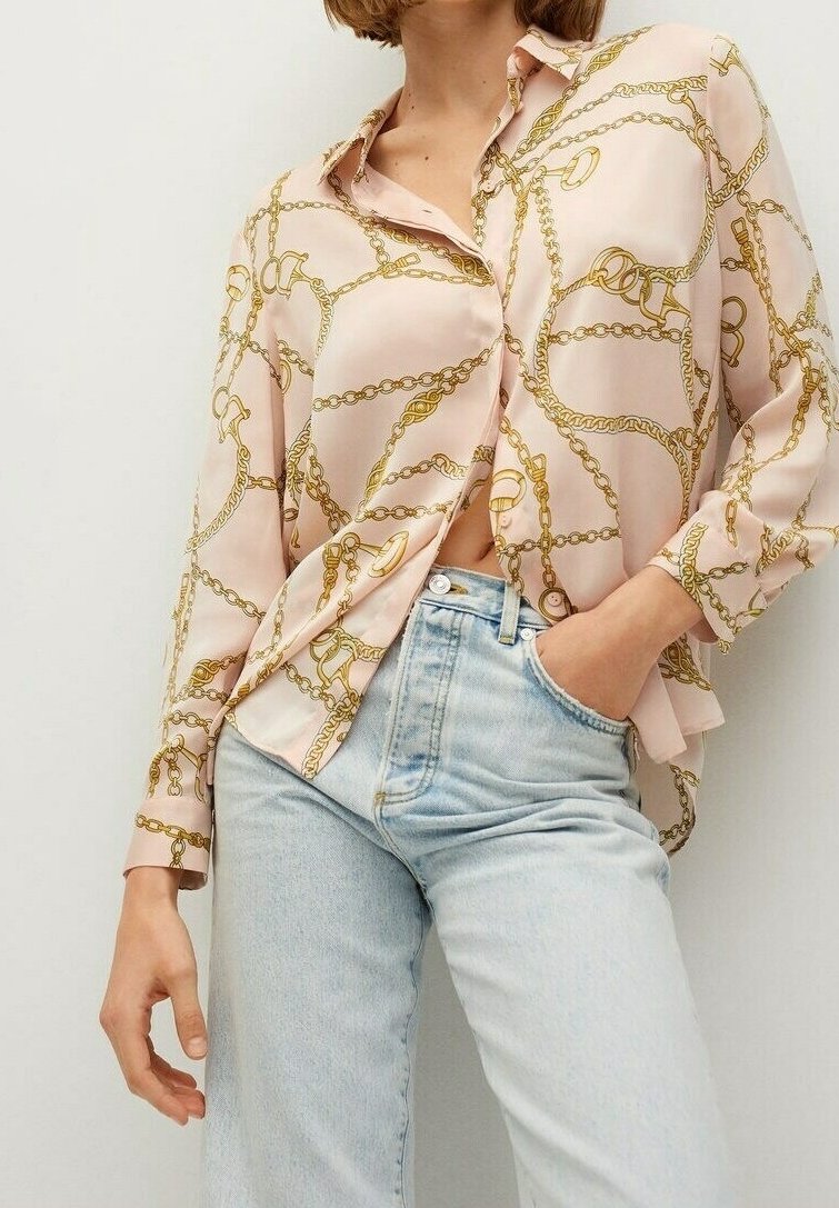 Personne portant une chemise rose pâle à boutons avec un imprimé chaîne dorée et un jean taille haute bleu clair, une main dans la poche et la chemise partiellement ouverte.