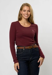 Cămașă ajustată cu mânecă lungă, de culoare vișinie, cu decolteu rotund și textură ribbed. Este asortată cu blugi închiși la culoare cu talie înaltă și o curea maro.