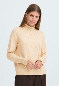 Pull en maille beige avec un col haut froncé, des motifs en losanges sur le devant et des poignets de manches froncés. Texture douce et coupe décontractée.