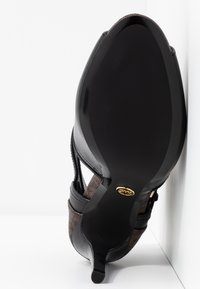 Semelle d'une chaussure à talon noire pour femme inclinée contre un mur blanc, présentant une semelle lisse et un petit emblème doré de la marque près du talon.