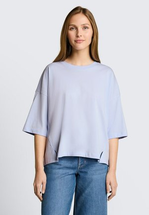 Jonge vrouw met steil haar die een los lichtblauw T-shirt met korte mouwen en middelblauwe spijkerbroek draagt, staand tegen een effen achtergrond.