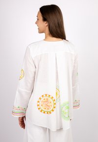 Weiße Linnenbluse mit langen Ärmeln, die bunte kreisförmige Muster, einen Rückenfalten und gestreifte Akzente an den Manschetten aufweist.