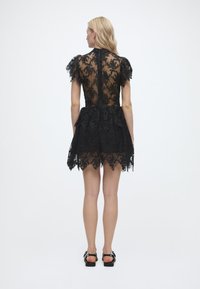 Γυναίκα που στέκεται με την πλάτη στραμμένη, φορώντας ένα κοντό μαύρο δαντελένιο φόρεμα με κοντά μανίκια και μαύρα επίπ εδα σανδάλια, σε λευκό φόντο.