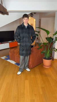Long plaid coat in dark gray with black, buttoned at the front. Lightly textured fabric, paired with gray trousers and white sneakers.