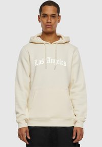 Kremfarget hettegenser laget av myk stoff med kengurulomme. Har hvit "Los Angeles" tekst i fet skrift. Elastiske mansjetter og kant.