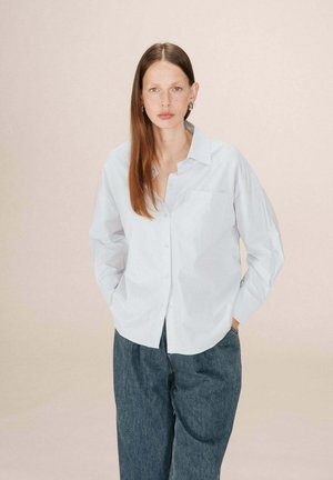 Chemise blanche à boutons avec col, manches longues et poche poitrine, assortie à un pantalon en denim sombre et ample, sur un fond doux.