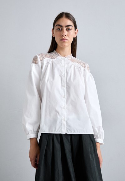 White blouse with lace shoulder detail, button front, and puffed sleeves. The fabric is smooth, paired with a dark, pleated skirt.