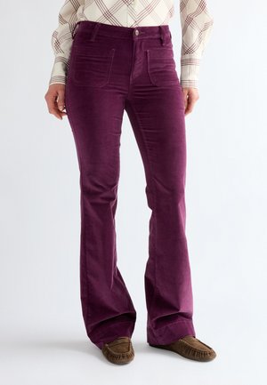 Donna che indossa pantaloni di velluto a coste viola a vita alta con tasche anteriori, abbinati a una camicia a quadri beige e mocassini marroni.