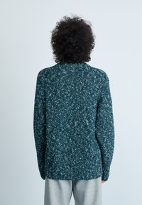 Gebreide trui in donker teal met een witte spikkeltextuur, een hoge kraag en lange mouwen. Het ontwerp heeft een relaxte pasvorm en een geribbelde zoom.