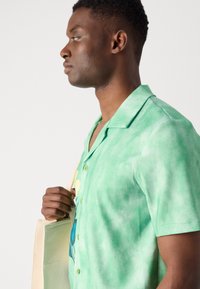 Grøn kortærmet skjorte med en blød tekstur, som har et tie-dye mønster og en enkelt lomme, båret med en beige tote taske.