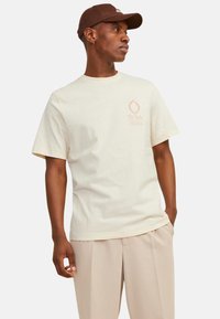 Cremefarvet bomulds t-shirt med korte ærmer, der har et trykt design i lys orange på venstre bryst. Bæres med beige bukser.