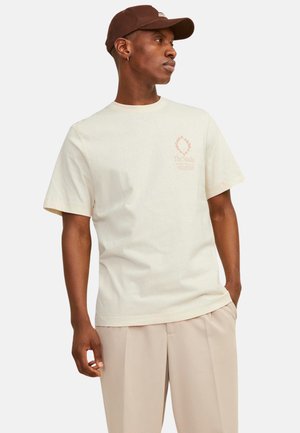 Krämbelädd bomullströja med korta ärmar, med en tryckt design i ljus orange på vänster bröst. Bärs med beige byxor.