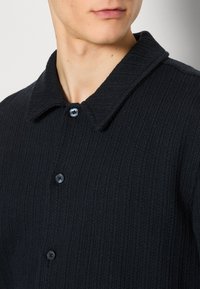 Schwarzer Strickcardigan mit strukturierter Oberfläche, ausgestattet mit einem Kragen und zwei dunklen blauen Knöpfen als Verschluss.