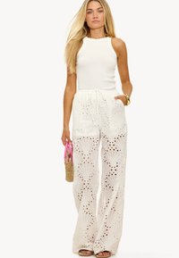 Witte mouwloze top gecombineerd met witte brede broek met ingewikkelde oogjes en uitsparingen. Accessoires omvatten een kleine geweven tas.