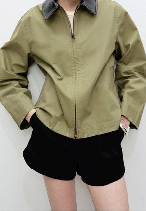 Osoba v olivově zelené bundě s černým límcem a černých šortkách, s rukama v kapsách, stojící před světlým neutrálním pozadím.