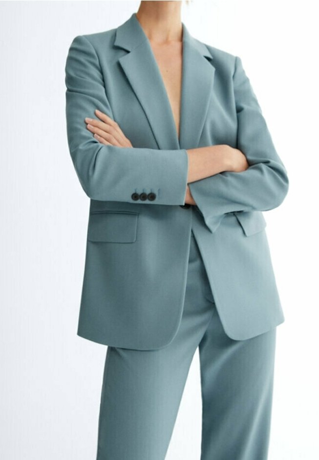 Personne portant un costume bleu sarcelle, les bras croisés, montrant un blazer avec des boutons sur la manche et un pantalon assorti.