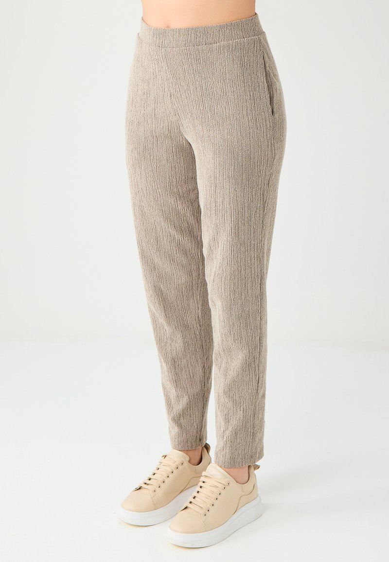 Beżowe, teksturowane spodnie w tapered z elastycznym paskiem w talii i bocznymi kieszeniami, w zestawieniu z jasnobeżowymi sneakersami na białym tle.