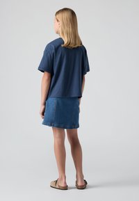 Mornarska majica kratkih rukava opuštenog kroja, u kombinaciji s plavom traper mini suknjom. Model nosi bež cipele bez kopčanja. Glatka tekstura, jednostavan dizajn.