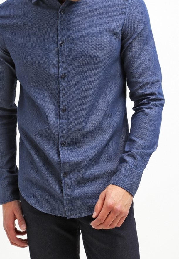 Homme portant une chemise texturée bleu foncé à manches longues et boutonnée, avec un pantalon noir, debout devant un fond blanc.