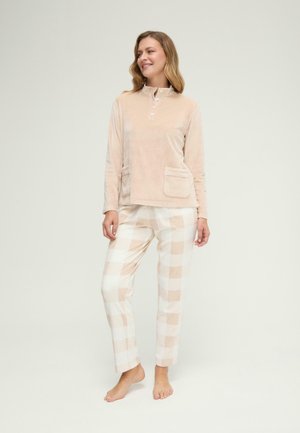 Mujer con camiseta beige de manga larga con bolsillos delanteros y pantalones beige y blancos de cuadros, de pie descalza frente a un fondo liso.