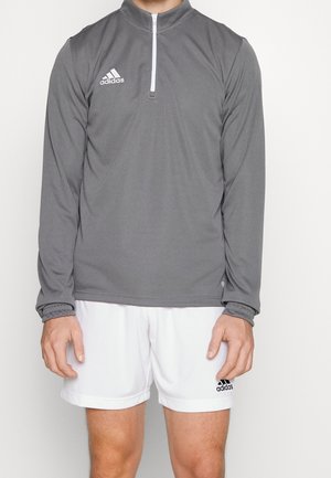 Mężczyzna ubrany w szarą bluzę Adidas z długim rękawem zapinaną na ćwiartkę oraz białe krótkie spodenki Adidas na jednolitym tle.