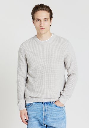 Giovane uomo con capelli castano chiaro, che indossa un maglione grigio chiaro a maglia e jeans blu, in piedi con una mano in tasca su uno sfondo bianco.