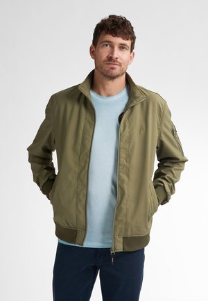 Olivgrüne Bomberjacke mit Reißverschlussöffnung, gerippten Bündchen und Kragen. Dazu ein hellblaues Hemd darunter, mit subtilen Logodetails auf der Brust.