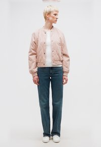 Άτομο με κοντά ξανθά μαλλιά, φοράει ανοιχτό ροζ γαζωτό μπουφάν, λευκό πουκάμισο, μπλε τζιν και λευκά αθλητικά παπούτσια, στέκεται και κοιτάζει προς τα πλάγια.