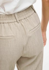 Pantalons beige clair avec une taille élastique et deux poches arrière. Le tissu a une texture fine avec un motif chiné subtil.