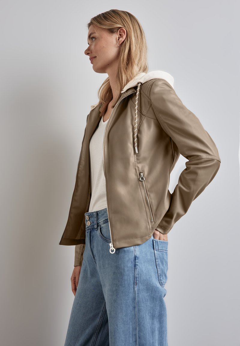 Giacca in ecopelle marrone chiaro con cappuccio bianco, chiusura con zip e tasche. Abbinata a jeans a gamba larga blu chiaro.