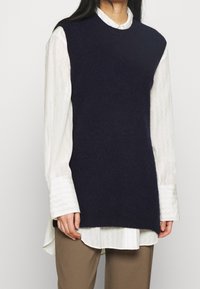 Marinblå stickad väst med rund halsringning, lager på lager över en vit strukturerad skjorta. Har en avslappnad passform och ribbade kanter.