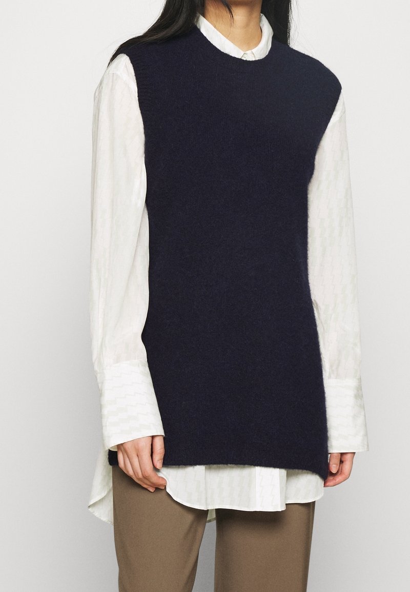 Marinblå stickad väst med rund halsringning, lager på lager över en vit strukturerad skjorta. Har en avslappnad passform och ribbade kanter.