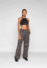 Sieviete stāv, tērpusies melnā sporta krūtīte, pelēkajos rakstainajos platā kāju biksēs ar auklu un melnās sporta apavās, rokas ieliktas kabatās.