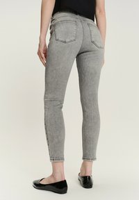 Osoba ubrana w jasnoszare, wysokiej talii dopasowane dżinsy i czarne lakierowane baleriny, stojąca tyłem do kamery.