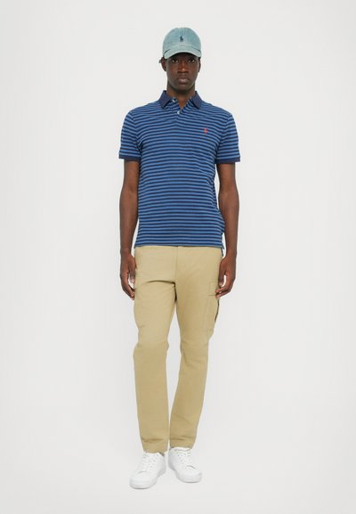 Jūras spēku svītrains polo krekls, beigo cargo bikses un baltas kedas. Cepure ir gaiši zila džinsa ar logo. Modelis stāv uz baltā fona.