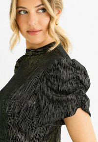 Black dress with gold metallic stripe pattern, high neckline, and puff sleeves. Soft texture and structured fit. Accentuated waist detail.