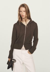 Brown zip-up hoodie made of textured knit fabric, featuring a hood, front zip closure, and ribbed hem. Paired with white pants and black bag.
