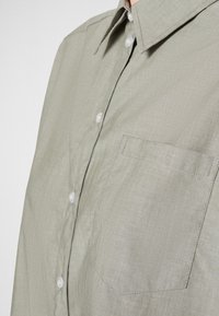 Camicia a bottoni verde chiaro con taschino sul petto, colletto a punta e texture sottile. Bottoni in plastica e dettagli di cucitura ordinati.