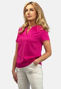 Kr ótki rękaw, wysoka góra w jaskraworóżowym materiale z plisowanym detalem. Sparowana z jasnymi spodniami, model stoi z rękami w kieszeniach.