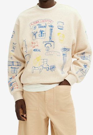 Beige sweatshirt med farverige abstrakte linjetegninger af bygninger, en person, planter og tekst, båret med matchende beige bukser.