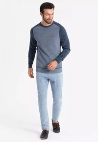 Sudadera azul clara con mangas y cuello de canalé en azul marino, cuerpo texturizado, pequeña etiqueta de logo en el pecho, combinada con jeans claros y zapatos oscuros.