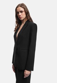 Sort skræddersyet blazer med dyb V-udskæring, strukturerede skuldre og lange ærmer. Har en enkelt knap og sidelommer for et minimalistisk design.