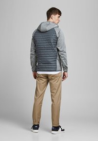Kapuz jacket mit einer grauen melierten Vorderseite und einem dunkelgrauen gesteppten Rücken. Kombiniert mit beigen Hosen und dunklen Sneakers.