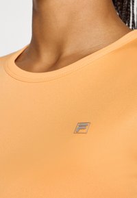 Nærbilde av en oransje sportsskjorte med en liten logo på brystområdet, som viser en jevn halslinje og stofftekstur.