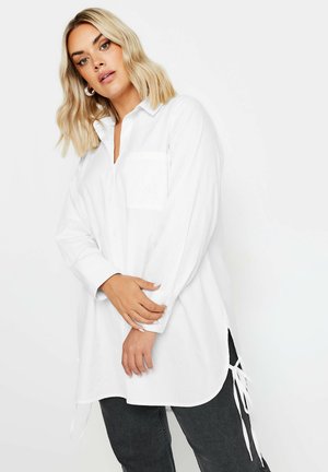 Camicia bianca con colletto a punte, maniche lunghe, tasca singola sul petto, spacchi laterali con lacci e vestibilità comoda. Tessuto di cotone liscio.