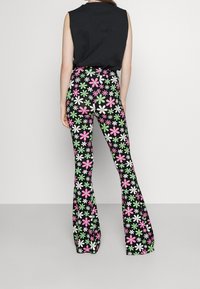 Vrouw in een zwarte mouwloze top en hoge zwarte flared broeken met een roze, groene en witte bloemenpatroon, gezien van achteren.