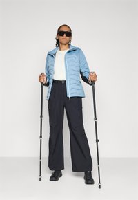 Lichtblauwe puffy jas met een capuchon over een witte top, gecombineerd met zwarte cargobroeken en zwarte schoenen. Houdt trekkingstokken vast en draagt een zonnebril.