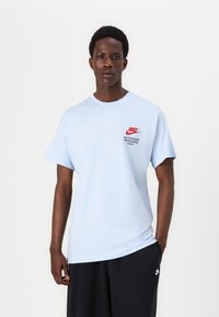 Gaiši zilā krāsā t-krekls, kas izgatavots no kokvilnas, ar sarkano Nike logotipu un tekstu "TIKAI AUTORIZĒTIE DARBA ŅĒMĒJI" uz krūtīm. Īsas piedurknes, apaļš kakla izgriezums.
